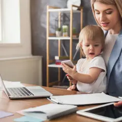 Kobieta z dzieckiem przy biurku z laptopem i tabletami. Czy na urlopie macierzyńskim można pracować na umowę zlecenie?