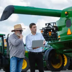 Dwóch rolników dyskutuje przy kombajnie. Jeden pokazuje coś na laptopie, drugi zastanawia się nad opłatą adiacencką a darowizną.