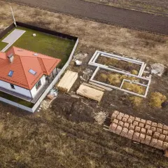 Działka zagrodowa w budowie: gotowy dom z czerwonym dachem, fundamenty nowego budynku, drewno i palety cegieł.