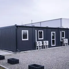 Szary kontener jako budynek gospodarczy, z oknami i klimatyzacją, stoi na żwirowym podwórzu.