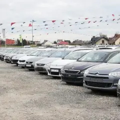 Rząd samochodów na placu, gotowych do sprzedaży. W tle domy i flagi. Możliwość zawarcia umowy komisowej samochodu.