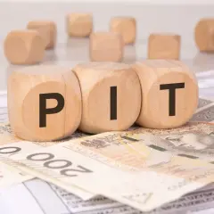 Drewniane klocki z literami "PIT" leżą na banknotach i dokumentach. Może to symbolizować rozliczenie podatkowe lub umowę darowizny.