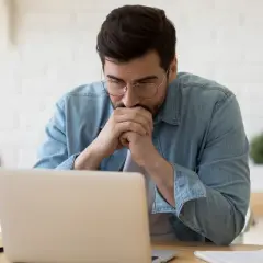 Mężczyzna w okularach pochyla się nad laptopem, zamyślony nad dokumentami, być może nad wnioskiem o odpis stanu cywilnego.