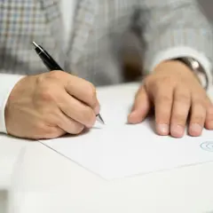 Mężczyzna w eleganckim garniturze podpisuje umowę komisową. Dokument z pieczęcią leży na białym biurku.