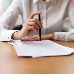 Dłoń trzymająca długopis nad stosem dokumentów, być może odpis aktu zgonu.