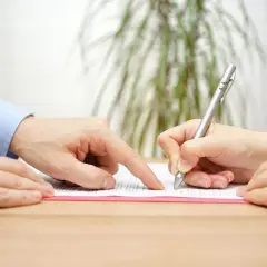 Mężczyzna wskazuje palcem na dokument, podczas gdy kobieta podpisuje go długopisem. Być może jest poręczycielem lub świadkiem.