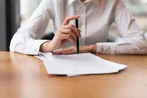 Dłoń trzymająca długopis nad stosem dokumentów, być może odpis aktu zgonu.