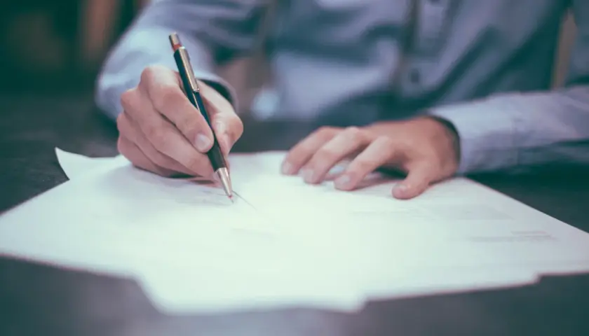 Dłoń pisze piórem na dokumencie. Czy komornik może zająć umowę zlecenie? To pytanie nurtuje wielu pracujących.