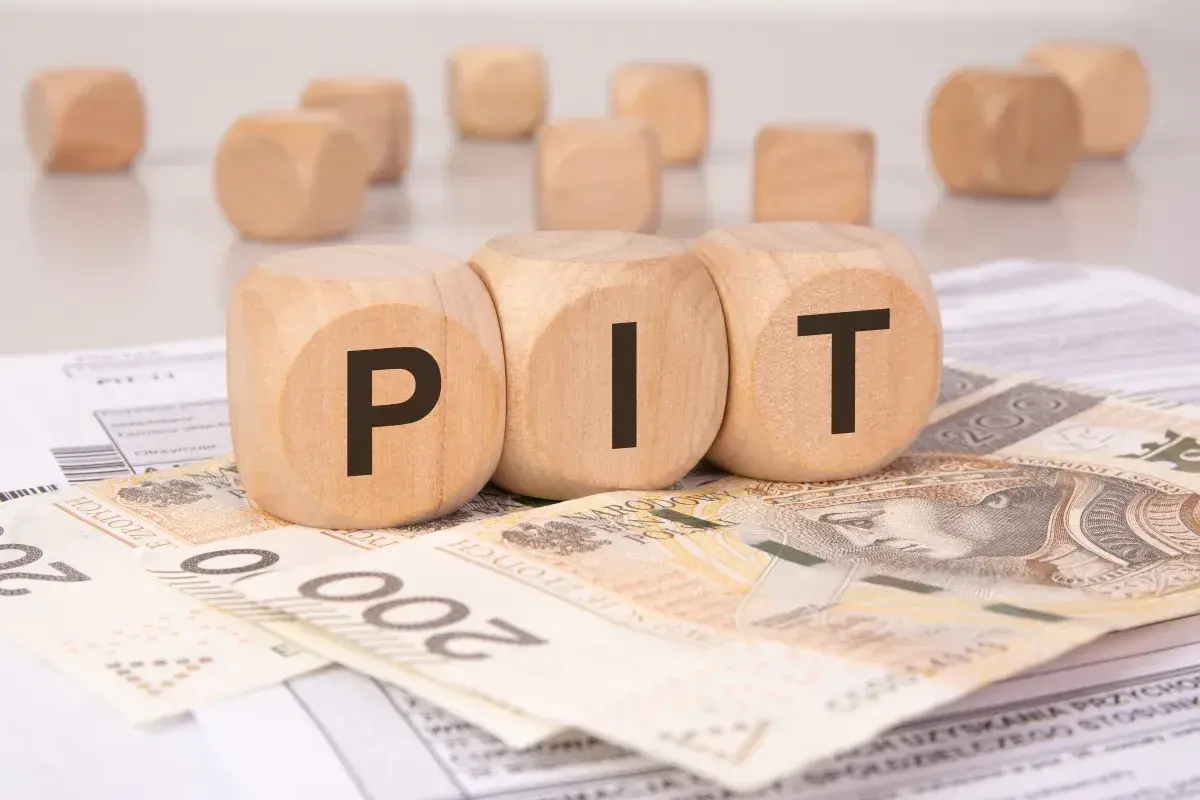 Drewniane klocki z literami "PIT" leżą na banknotach i dokumentach. Może to symbolizować rozliczenie podatkowe lub umowę darowizny.