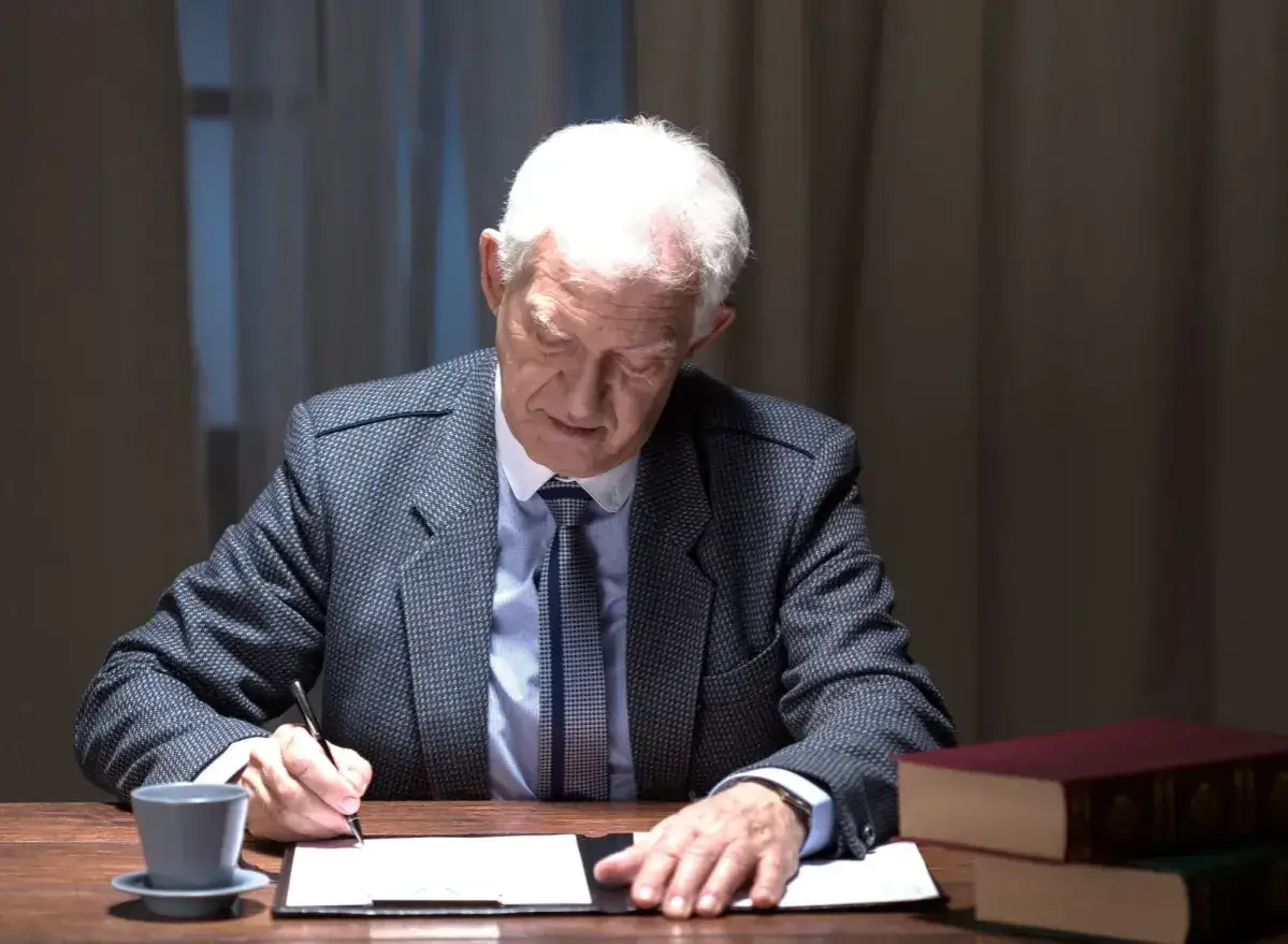 Starszy mężczyzna w garniturze podpisuje dokumenty. Zastanawia się, ile kosztuje akt zgonu, gdy obok leżą grube księgi.