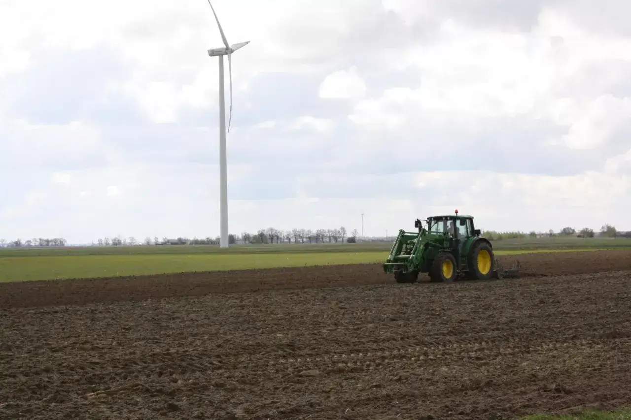 Zielony traktor orze pole, a w tle widać wiatrak. To obraz rolnictwa, gdzie dzierżawa a zasiedzenie to ważne kwestie.