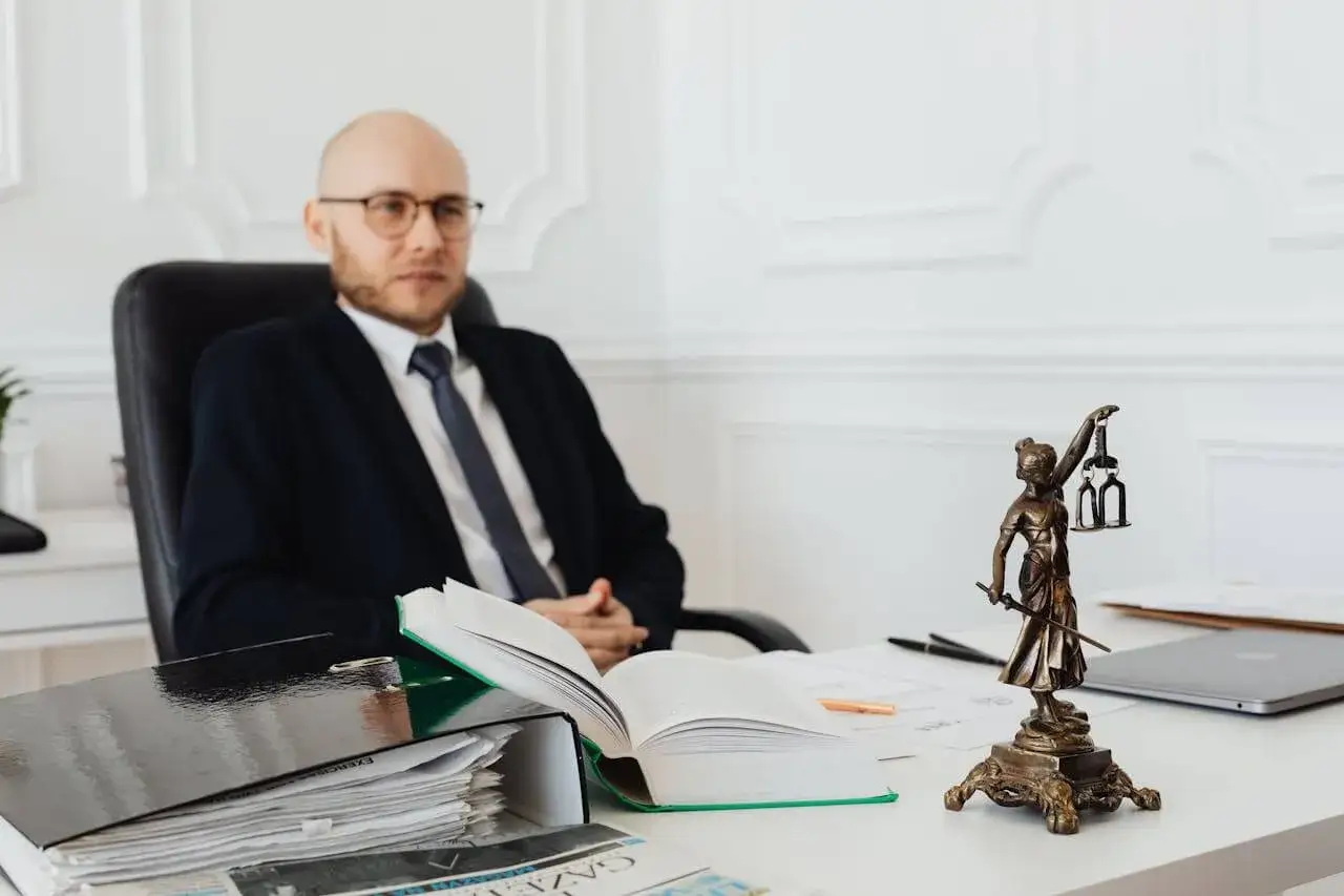 Prawnik w okularach siedzi przy biurku, na którym leży otwarta księga i dokumenty. Obok stoi figurka Temidy. Dowiedz się, ile kosztuje wpis do księgi wieczystej u notariusza.