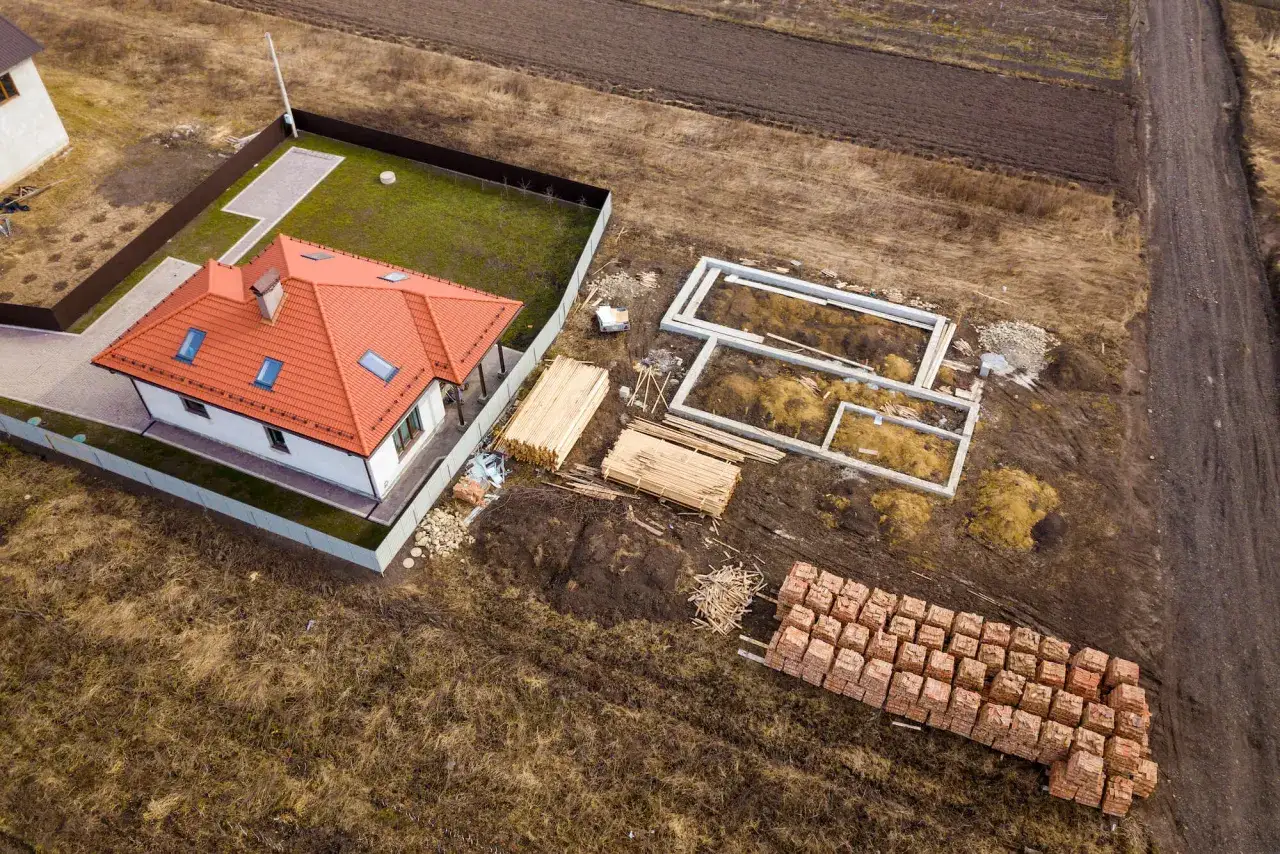 Działka budowlana z gotowym domem i fundamentami pod kolejny. Materiały budowlane wskazują na planowany podział nieruchomości na powiększenie działki sąsiedniej.